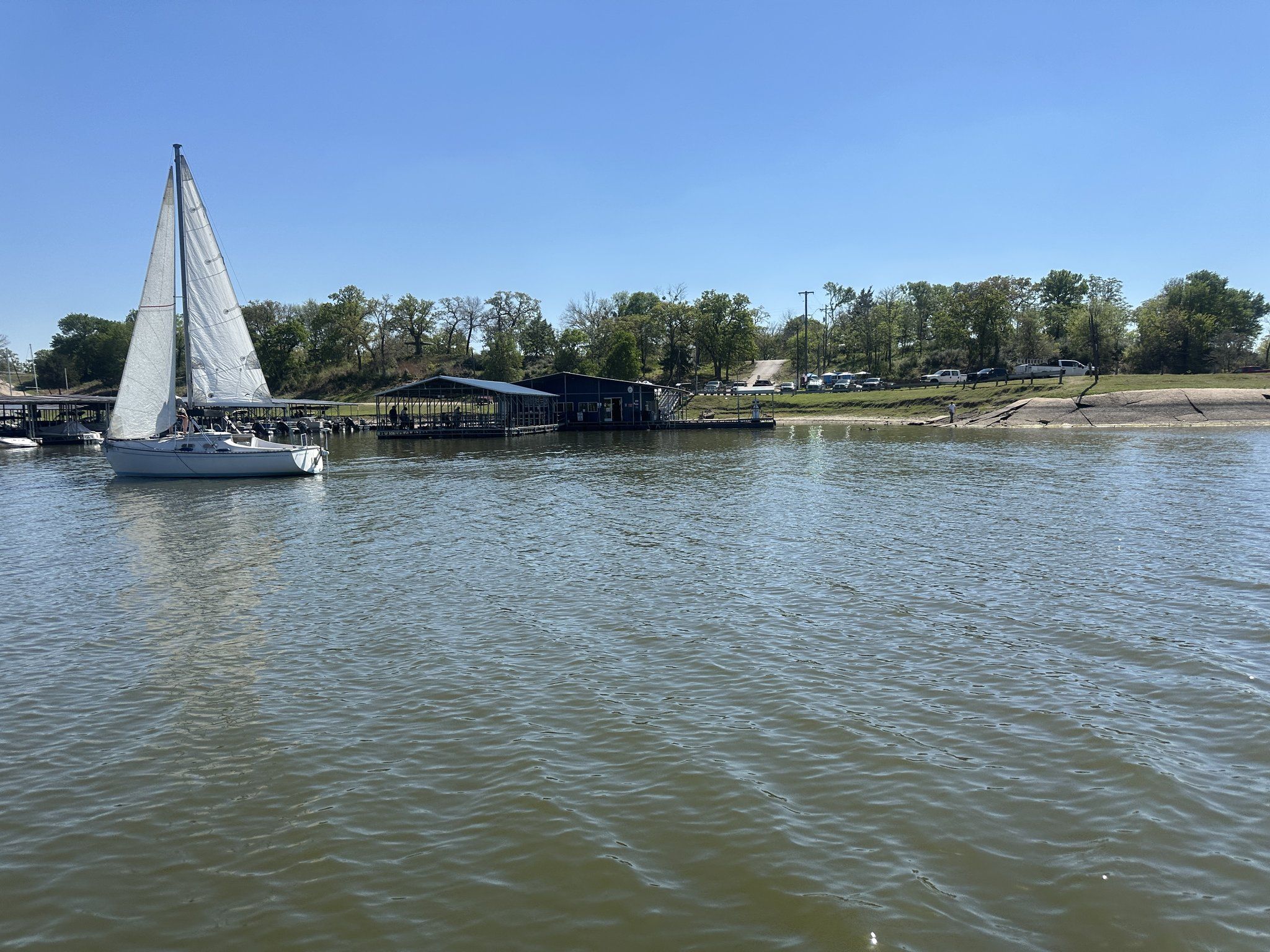 A white sailboat passing in front of the covered slips and wooded shoreline of the marina on Lake Somerville