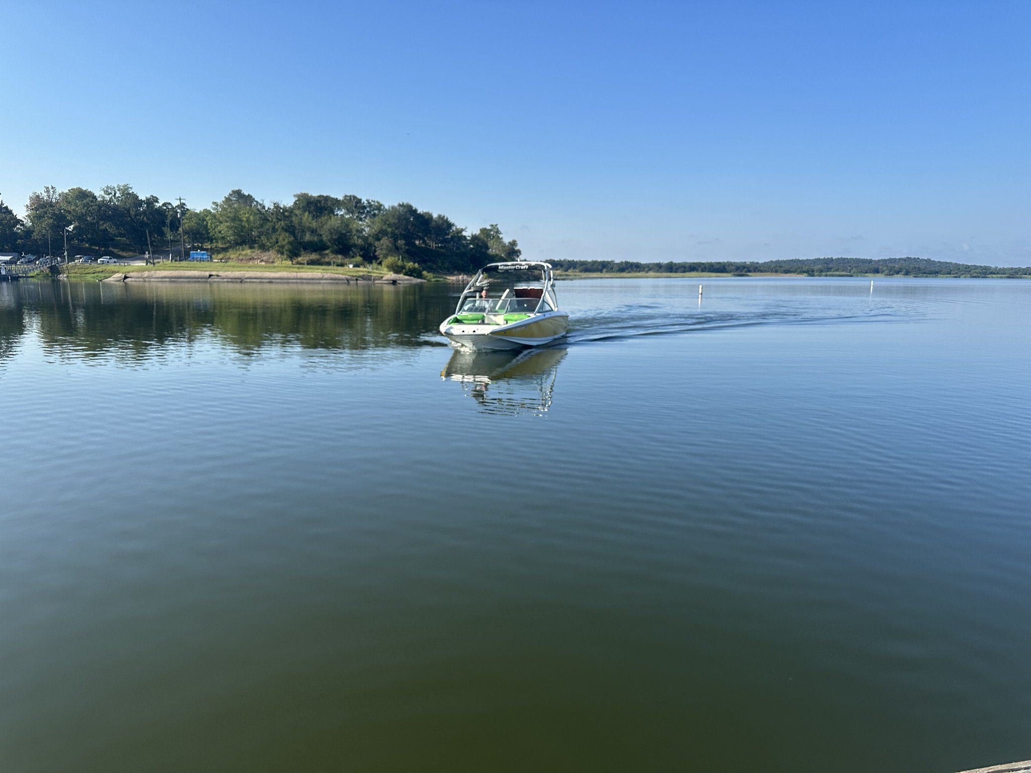 A white and green wakeboat sitting still on flat water that mirrors the wooded shoreline and blue sky