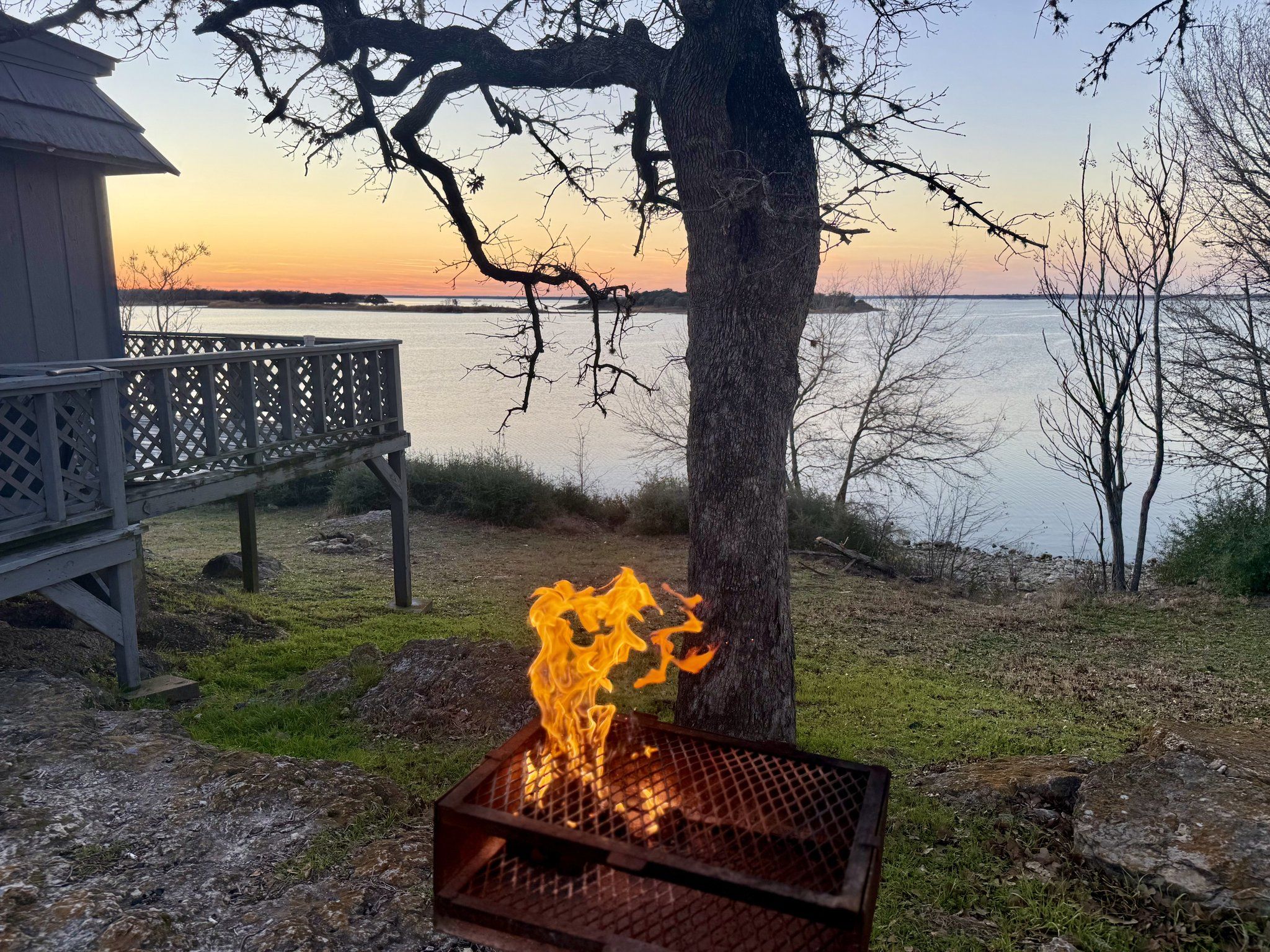Campfire burning in a steel ring on the bluff with camp chairs and the sun setting over Lake Somerville