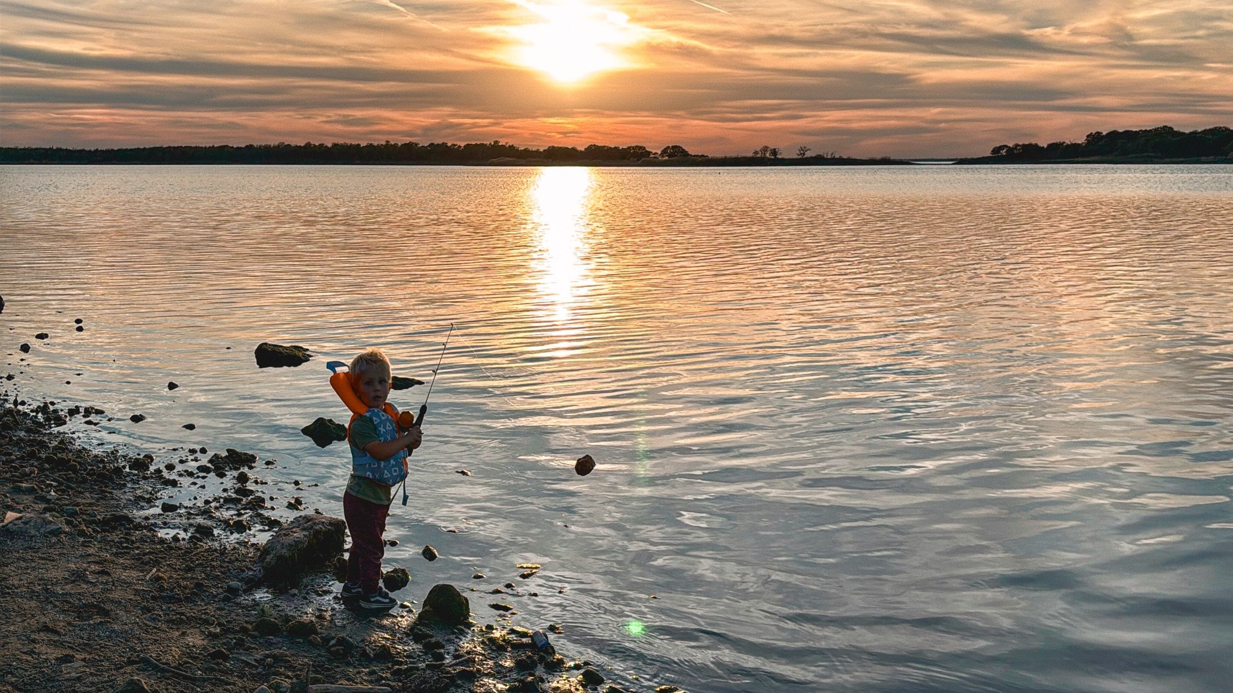 A small child in a life jacket fishing from the rocky shore of Lake Somerville at sunset