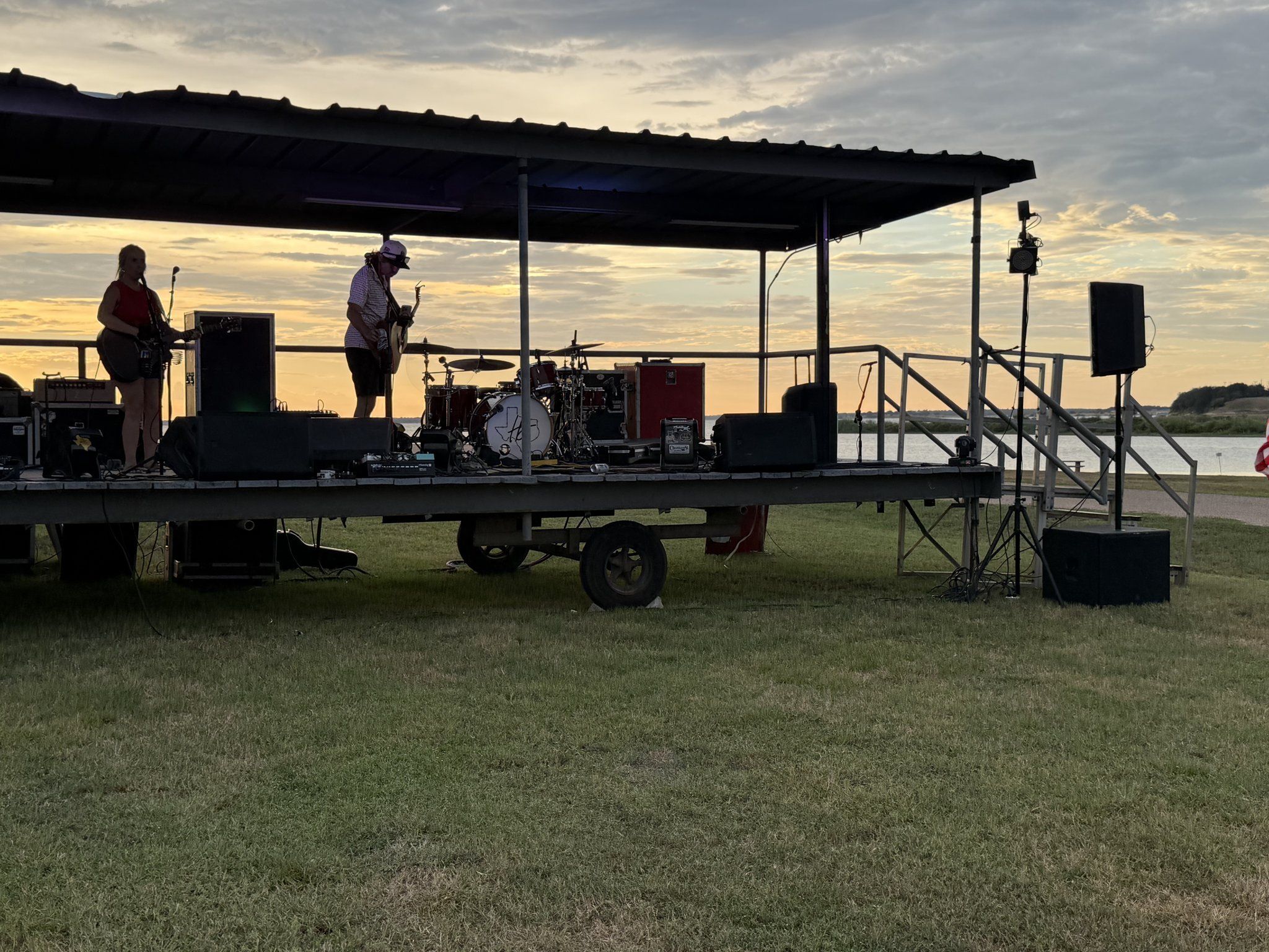 A band playing on the covered stage with a drum kit at dusk, with Lake Somerville behind
