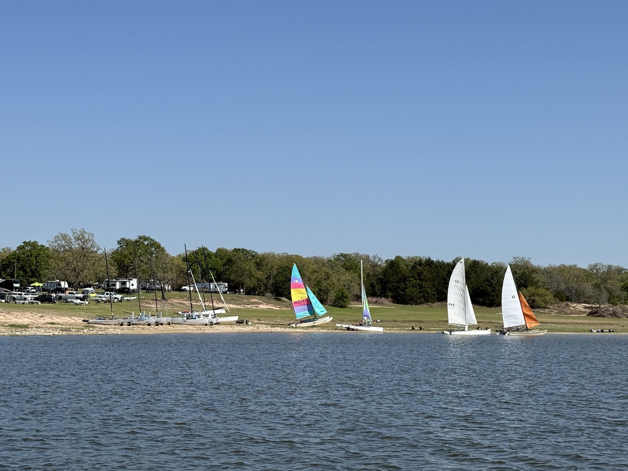 A line of catamarans and dinghies with brightly colored sails pulled up along the sandy shoreline, with RVs parked on the grass behind.