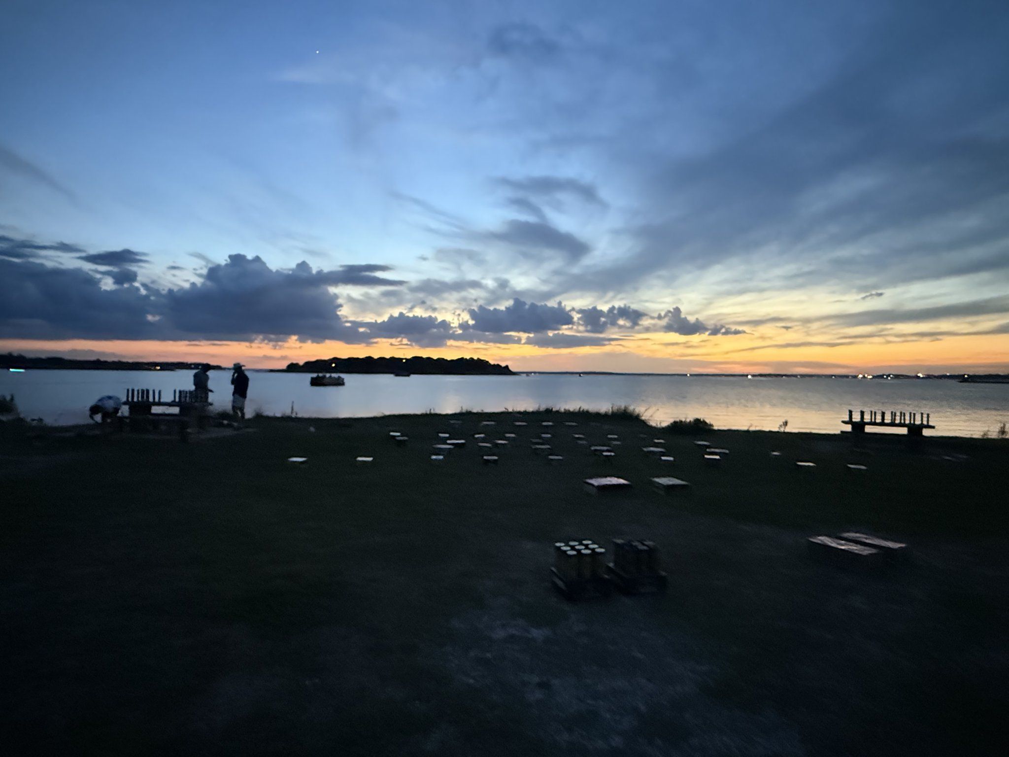 Rows of washer boards laid out across the lakefront lawn at dusk, with a couple of players at the far end under a blue and gold sky.