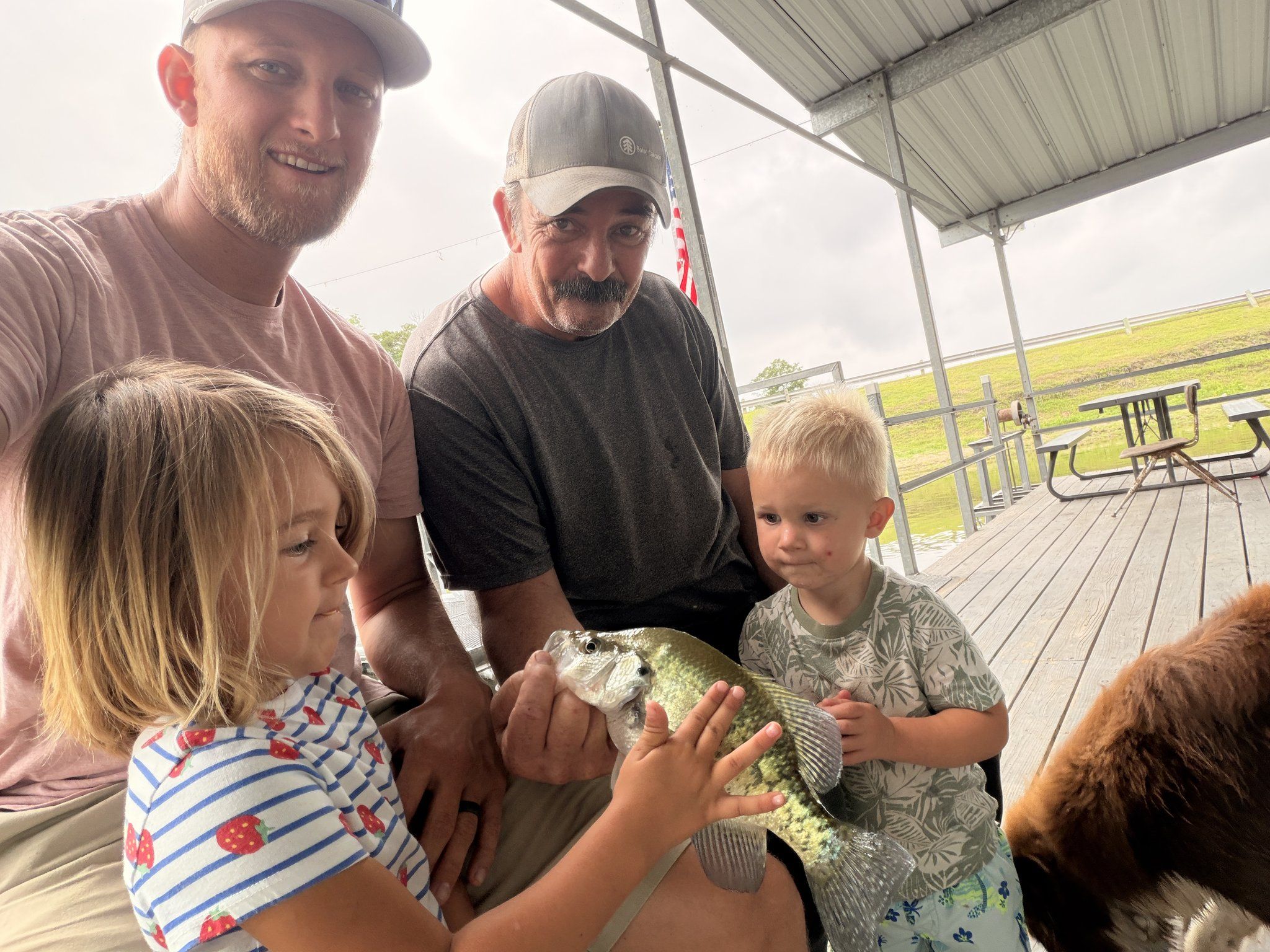 Two men crouch on the covered dock with two small children as they all hold a crappie together.