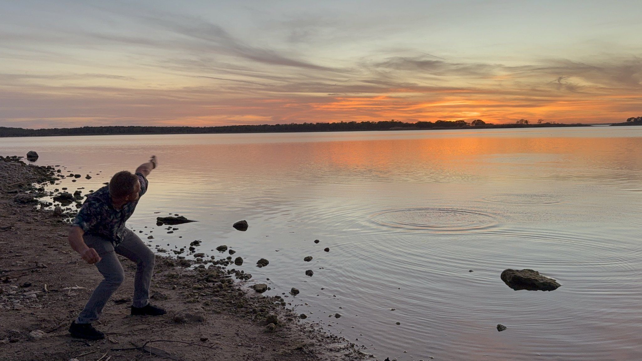A man mid-throw skipping a stone across glassy water at sunset, ripples spreading under a peach and orange sky.