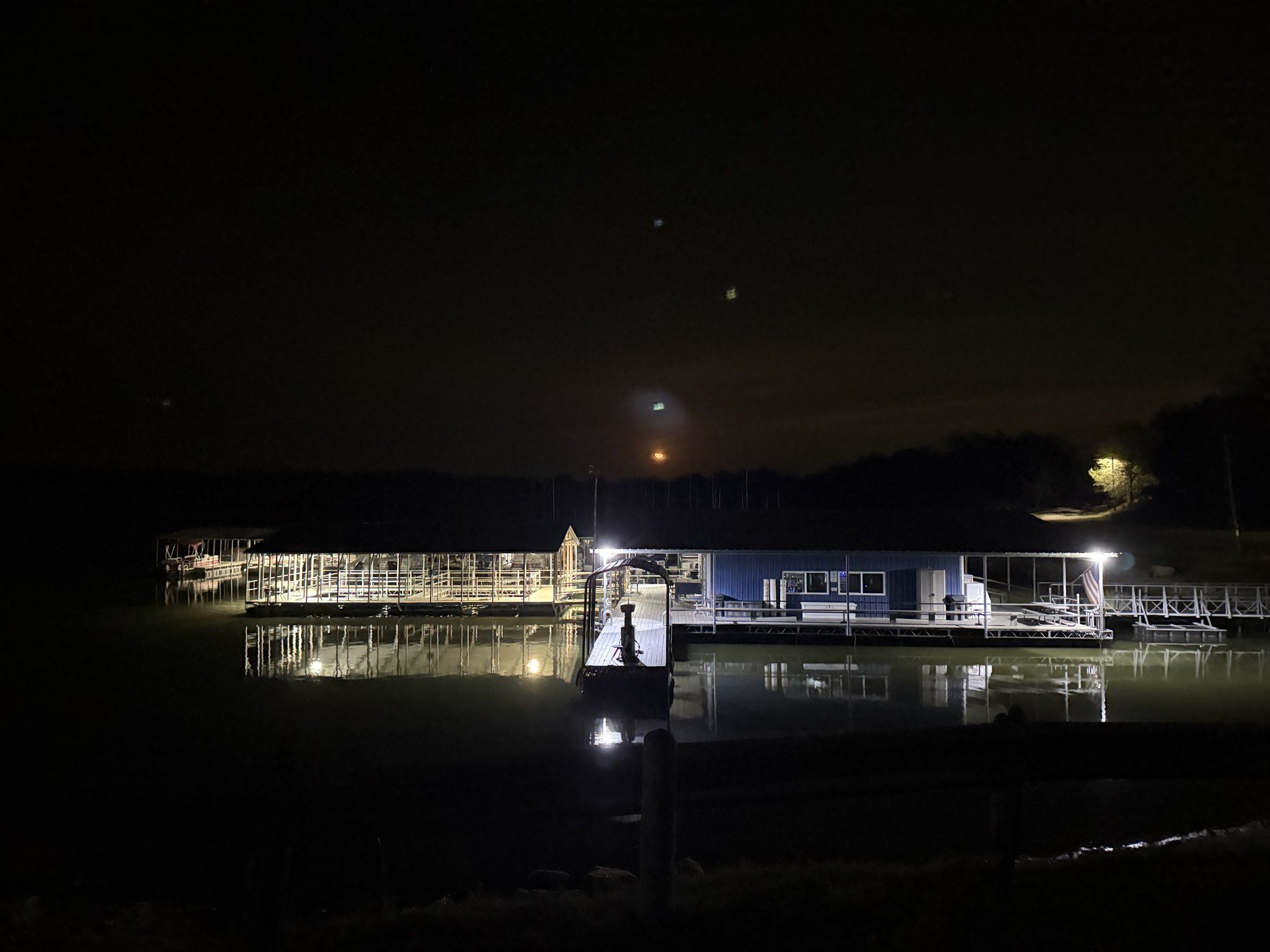 The blue marina building and covered docks lit by floodlights at night and mirrored in still water