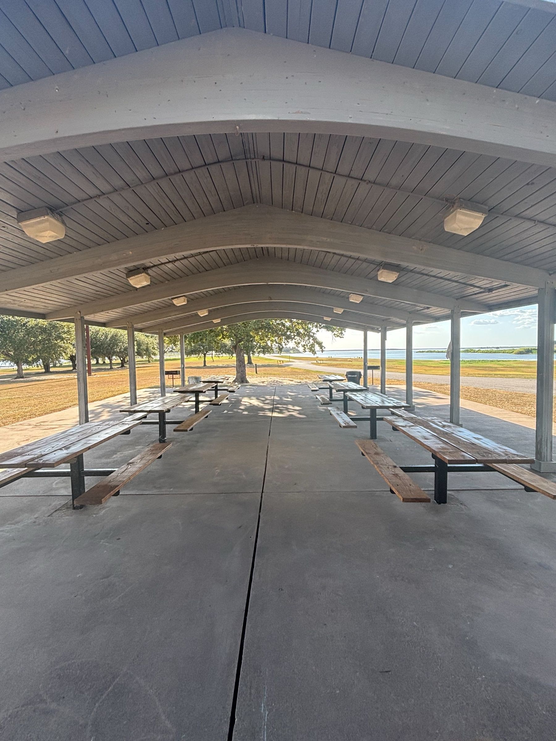 Looking down the length of the pavilion under its arched wooden roof, with picnic tables on both sides and lake and oak trees beyond.