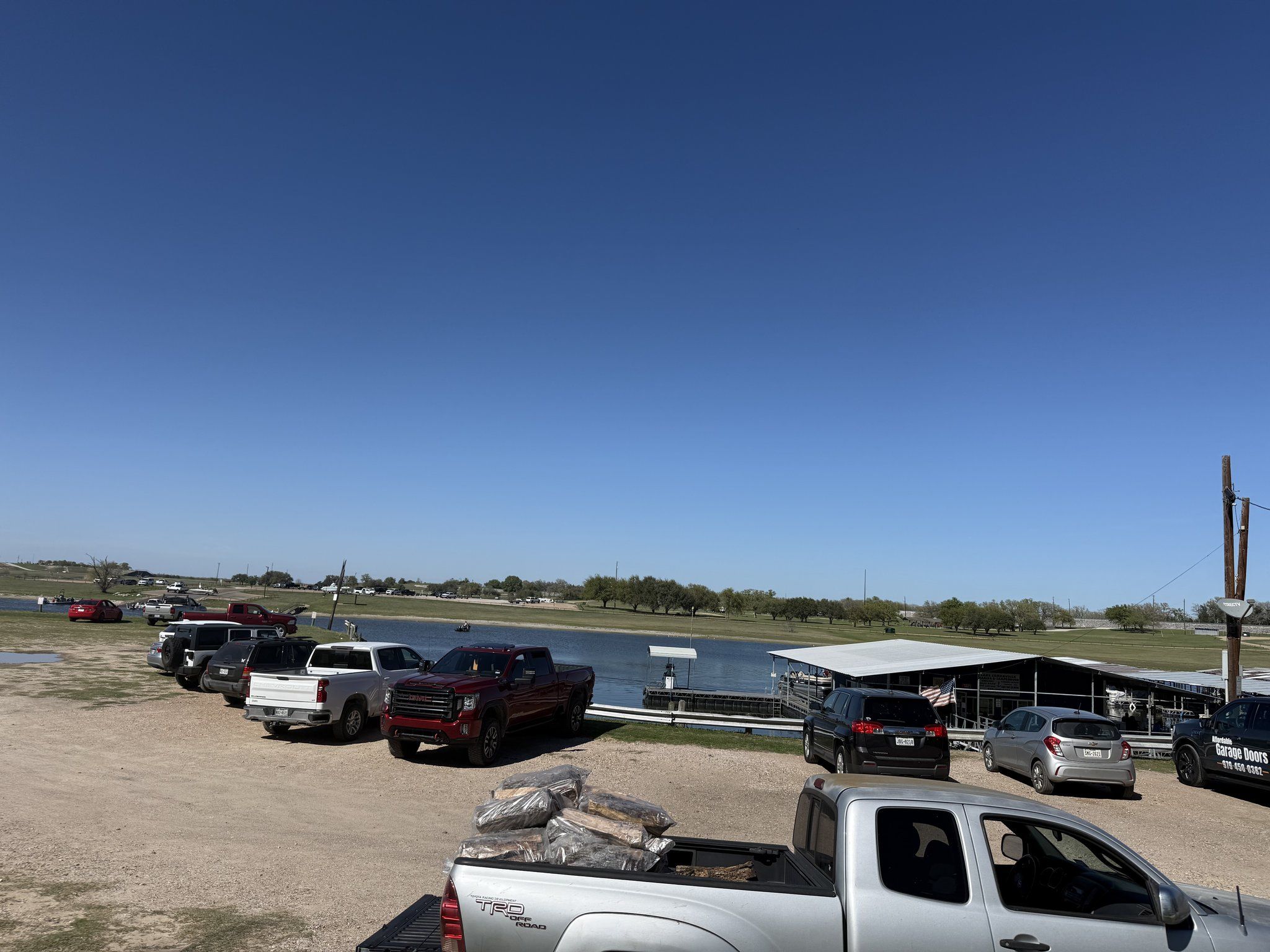 The gravel staging lot packed with pickup trucks and boat trailers on a bright day, with the covered slips and lake beyond.