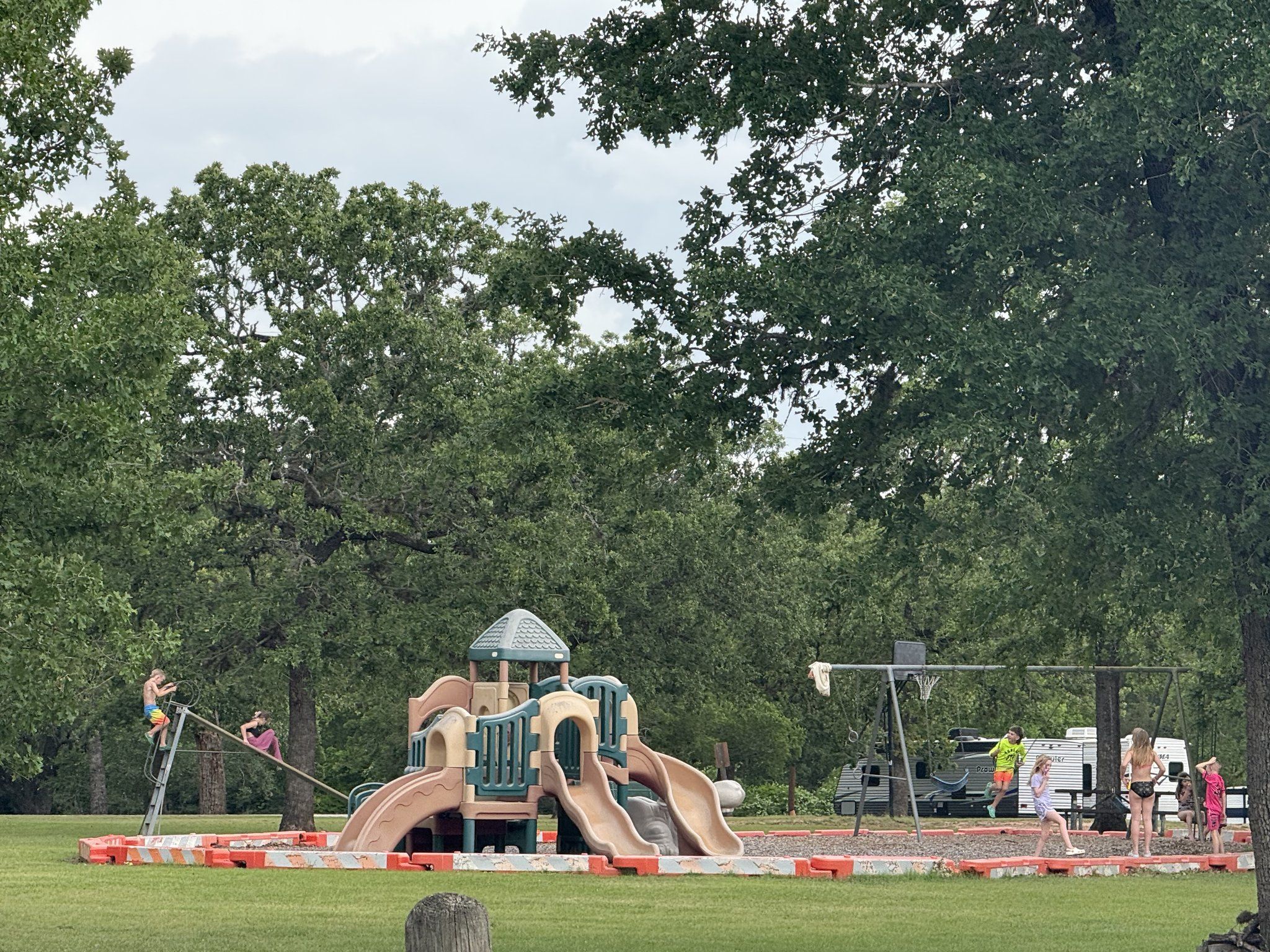 Children playing on a playground with slides, a climbing frame and swings among tall oaks