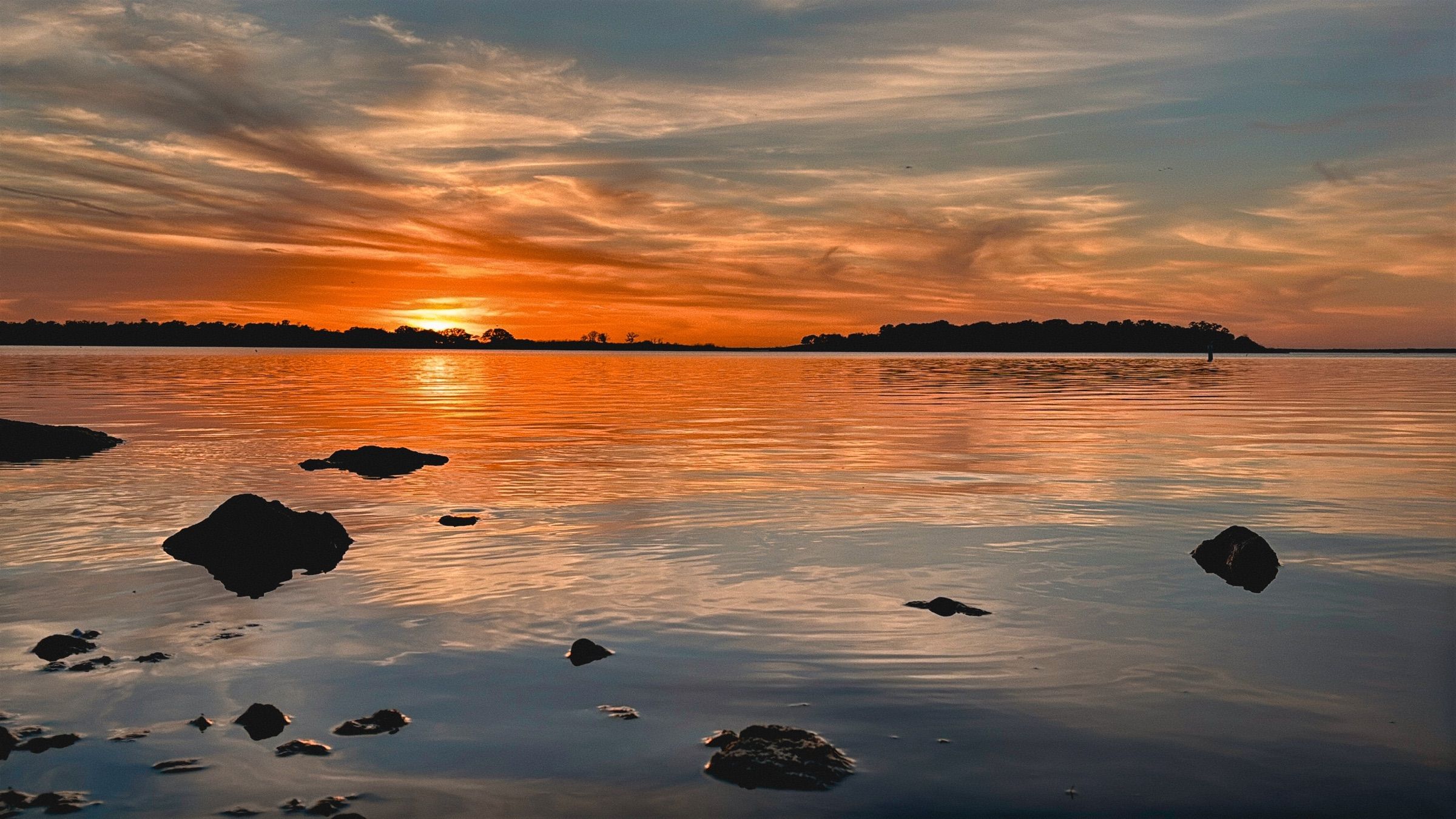 Orange sunset reflected in still water beside dark rocks on Lake Somerville